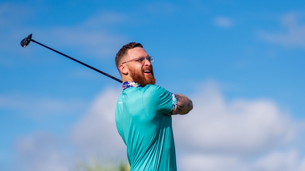 A man with a beard and glasses swings a golf club outdoors under a blue sky with clouds, wearing a teal shirt, pondering how long does golfing take.